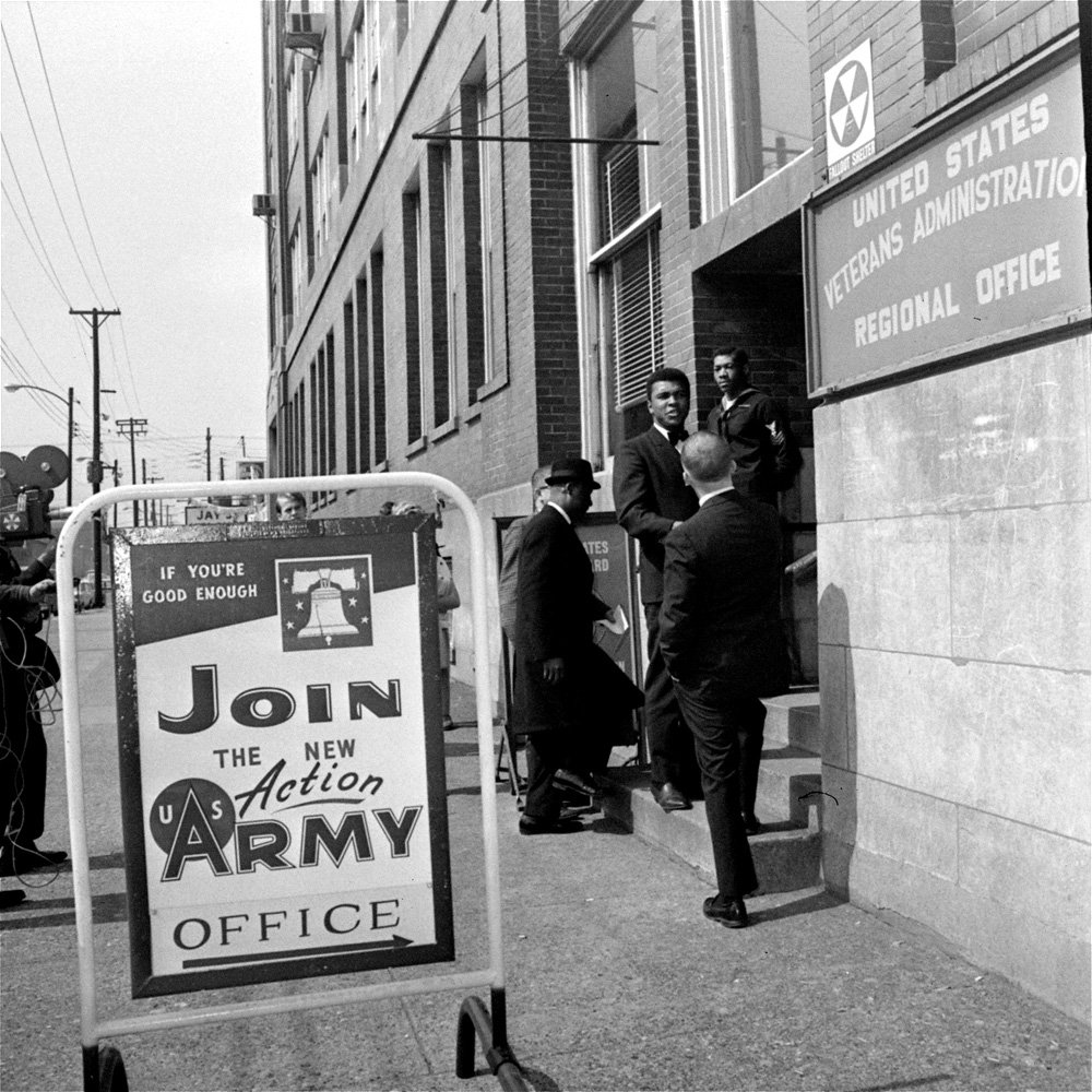 Heavyweight boxing champ Cassius Clay, paused on the step and greeted a friend, March 17, 1966, as he arrived at the Veterans building to appeal his 1A draft classification. Behind him, dark coat and hat, is his attorney, Edward Jocko of New York.  (AP Photo)